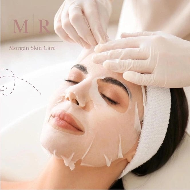 A woman lies on a spa bed with her eyes closed, wearing a white sheet mask on her face. A person wearing white gloves gently applies or adjusts the mask on her forehead.